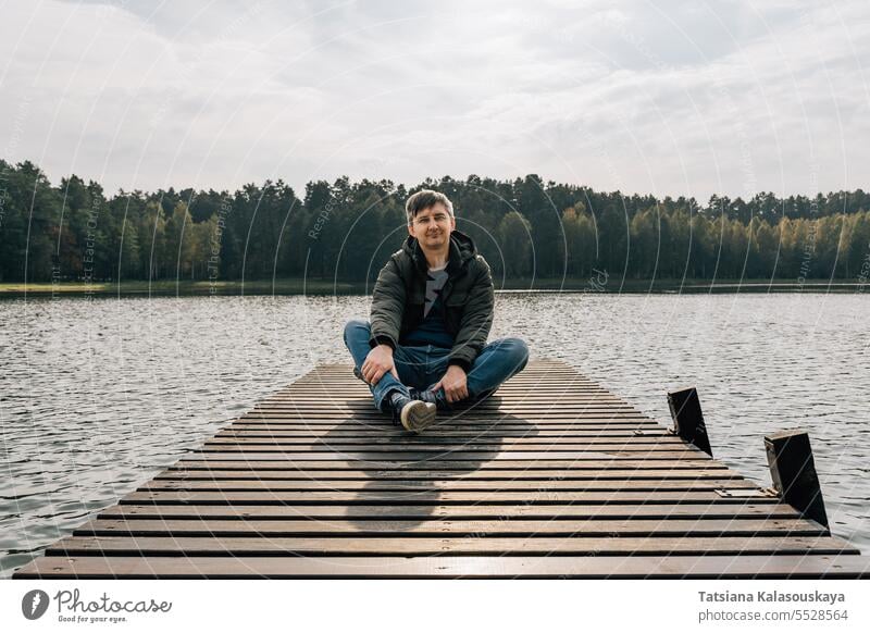 Ein Mann genießt einen sonnigen Herbsttag, während er auf einem Holzsteg an einem See sitzt Jeanshose Trägerkleid Jacke sitzen entspannend hölzern