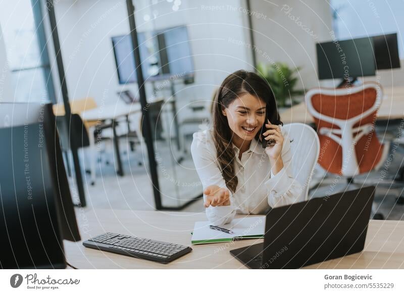 Junge Geschäftsfrau, die ein Mobiltelefon benutzt, während sie an einem Laptop in einem modernen Büro arbeitet Beruf Smartphone Frau jung Glück sprechend