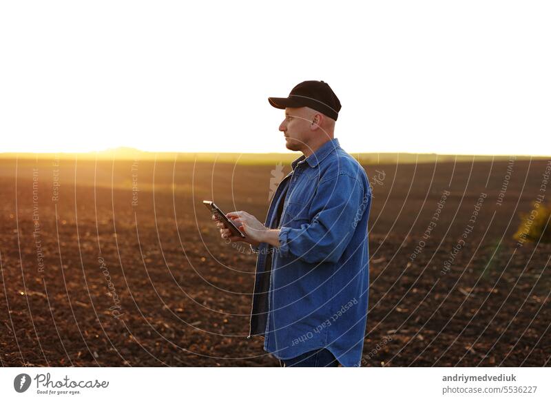 Intelligente Landtechnik und Landwirtschaft. Ein Landwirt benutzt ein digitales Tablet auf einem Feld mit gepflügtem Boden bei Sonnenuntergang. Überprüfung und Kontrolle der Bodenqualität, der Bereitschaft zur Aussaat von Pflanzen und zum Anbau von Gemüse