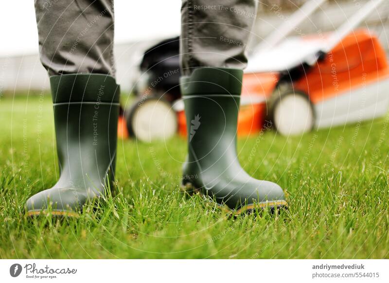 Abgeschnittenes Foto von männlichen Gärtner Beine in Gummistiefeln steht auf geschnittenem grünem Gras Rasen im Hinterhof des Hauses. Moderne elektrische schnurlose Rasenmäher auf der Rückseite. Landscaping Industrie Thema.