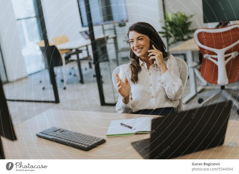 Junge Geschäftsfrau, die ein Mobiltelefon benutzt, während sie an einem Laptop in einem modernen Büro arbeitet Beruf Smartphone Frau jung Glück sprechend