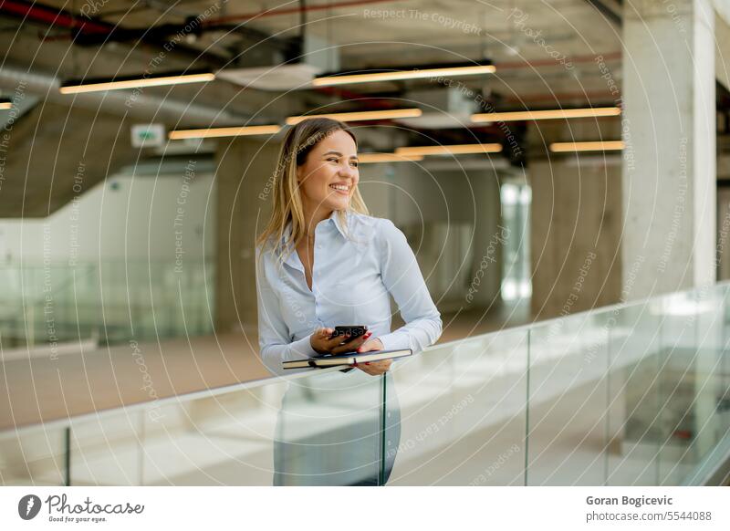 Junge Frau mit Notebook, die ein Mobiltelefon benutzt und in einem modernen Büroflur steht Erwachsener attraktiv schön Business Geschäftsfrau lässig Unternehmen