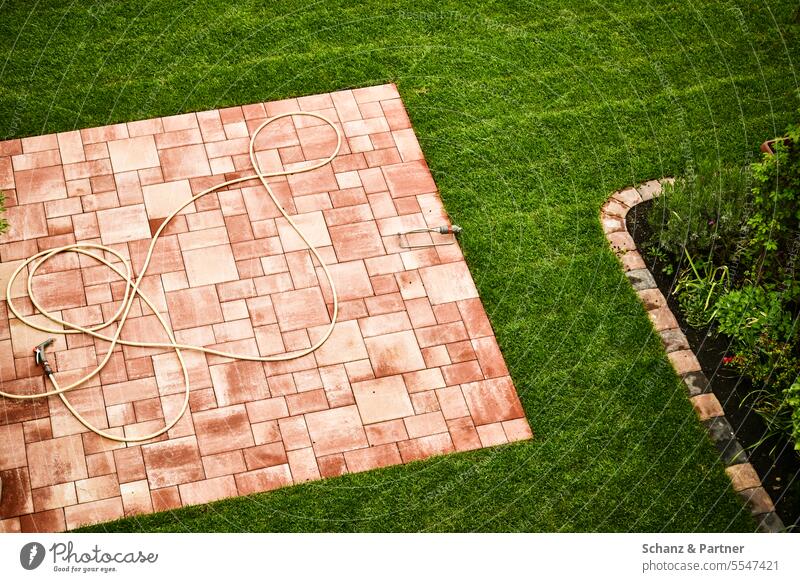 Gartenschlauch liegt auf einer quadratischen Terrasse aus Steinfliesen in einem gepflegten Garten mit Rasen und einem Pflanzstreifen bürgerlich ordentlich Wiese