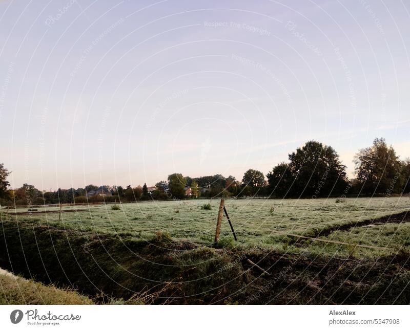 Morgenstimmung im Frühherbst, eine eingezäunte Weide mit Entwässerungsgraben im Vordergrund und Bäumen und Häusern hinter der Weide. früh morgens Stimmung