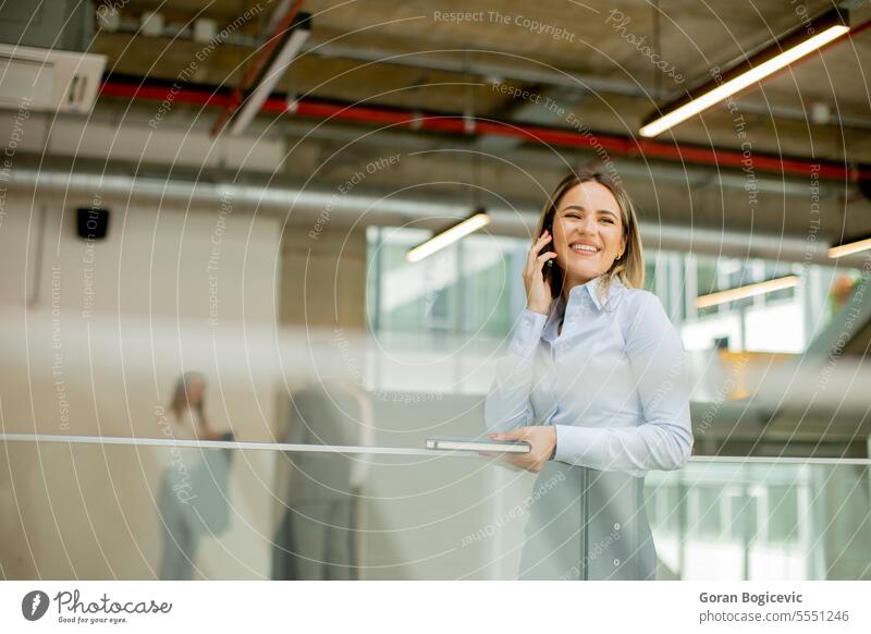 Junge Frau mit Notebook, die ein Mobiltelefon benutzt und in einem modernen Büroflur steht Erwachsener attraktiv schön Business Geschäftsfrau lässig Unternehmen