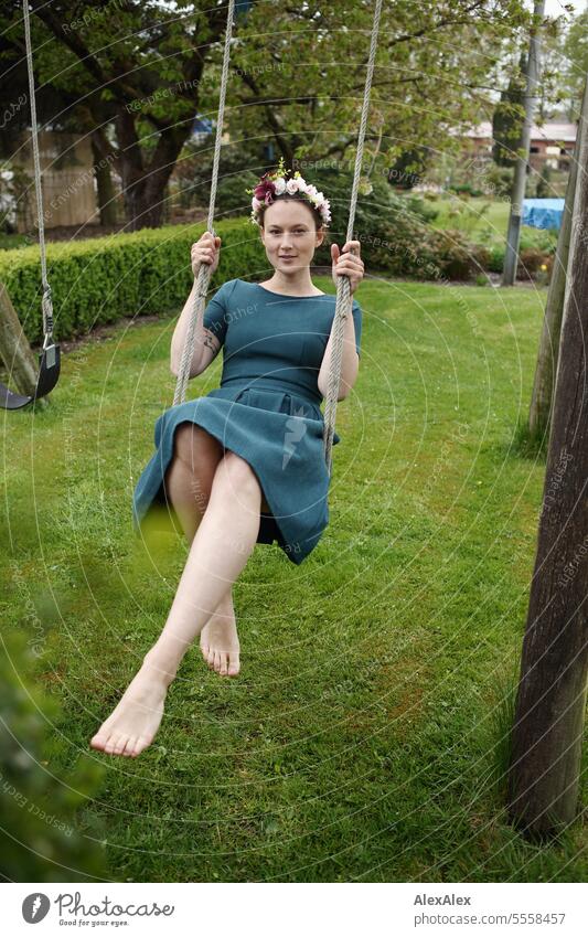 Portrait einer jungen Frau im Garten auf einer Schaukel - barfuß mit langen Beinen und Blumenkranz im Haar junge Frau Kleid schlank sportlich schön anziehend