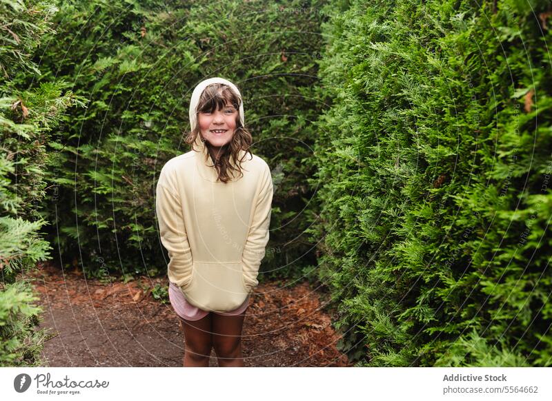 Lächelndes Mädchen mit Kapuzenpulli in der Nähe von grünen Bäumen Kind nadelhaltig Park Baum Straße Glück Landschaft stehen Hand-in-Tasche positiv Natur