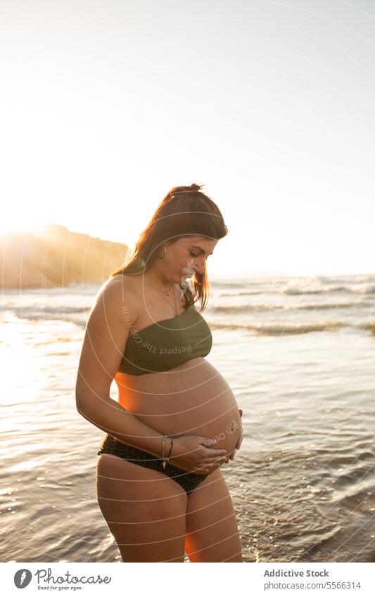 Schwangere Frau im Meerwasser stehend schwanger Bauch erwarten MEER Bikini Sommer Sonnenuntergang berühren Glück heiter Lächeln pränatal vorwegnehmen