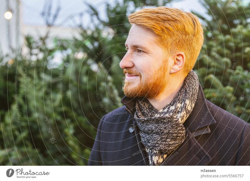 rothaariger mann schaut seitwärts unter weihnachtsbäumen auf einem straßenmarkt echtes Gelächter freudiger Ausdruck Mantel Lächeln Fröhlichkeit Blick überrascht