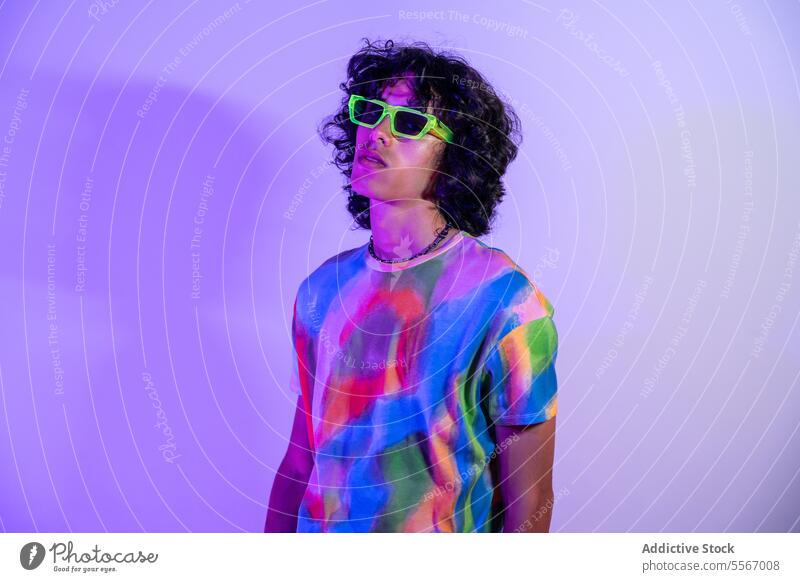 Lateinamerikanischer Mann mit buntem Hemd und grüner Sonnenbrille lockig Behaarung tie-dye Fliederbusch Hintergrund Mode Stil lässig Farbe pulsierend Pose Model