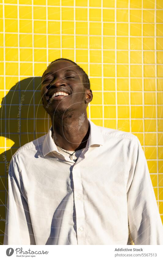 Junger Mann mit hellem Lachen jung freudig gelb Fliesen u. Kacheln Wand Sonnenlicht Schatten weiß Hemd pulsierend Glück heiter positiv Emotion Gesicht Zähne