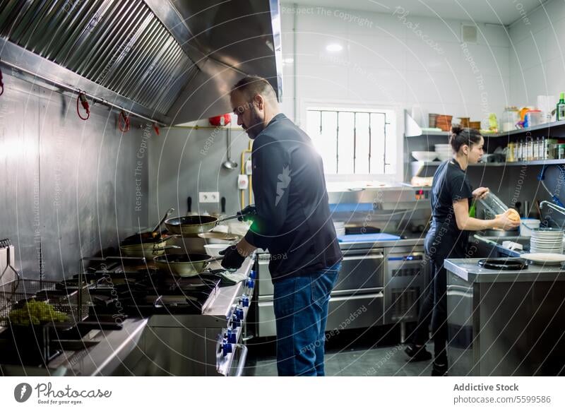 Seitenansicht eines bärtigen männlichen Kochs und einer Frau, die über einer Pfanne kochen und das Geschirr in einer modernen Restaurantküche waschen Küchenchef