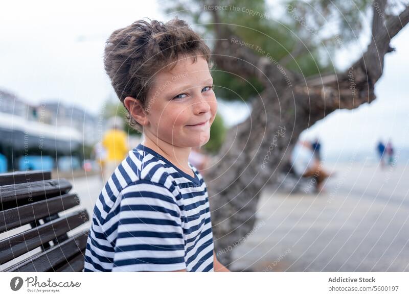 Ein Kind in einem gestreiften Hemd wirft einen spielerischen Blick über die Schulter in der Nähe einer baumbestandenen Promenade MEER gestreiftes Hemd Baum