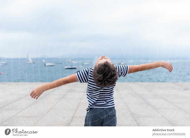 Kind mit ausgestreckten Armen steht am Meer und drückt Freiheit und Freude aus.Titel: Fröhliches Kind am Meer MEER Arme ausgestreckt gestreiftes Hemd Horizont