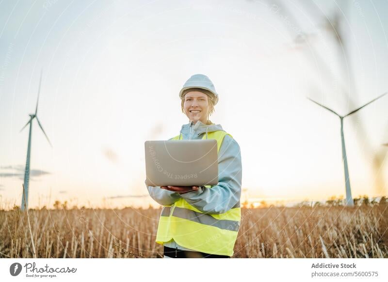 Ingenieur, der mit seinem Laptop im Windpark bei Sonnenuntergang in die Kamera schaut Frau Windkraftanlage Feld in die Kamera schauen Fangvorrichtung