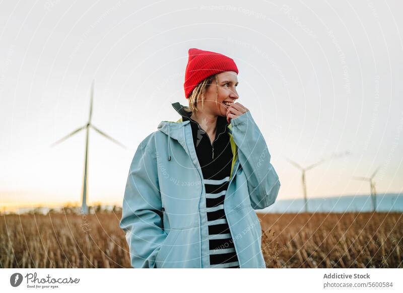 Lächelnde, alternative Frau in Jeansjacke, die von Windrädern wegschaut gelb Beanie Windkraftanlage nachhaltig Ökostrom Wegsehen heiter jung Erwachsener Porträt