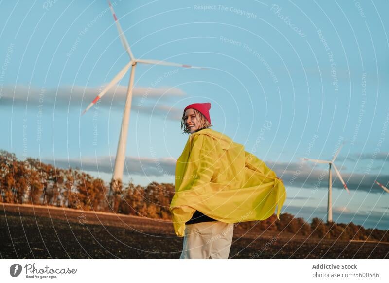 Fröhliche Person in Gelb in der Nähe von Windrädern bei Sonnenuntergang gelb Regenmantel rosa Beanie Lächeln Feld Windkraftanlage Himmel heiter