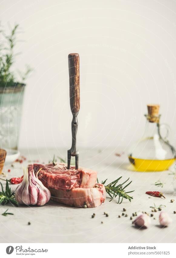 Rohes Fleischsteak mit Zutaten und Fleischgabel auf dem Küchentisch mit Rosmarin, Pfeffer und Olivenöl. Kochen zu Hause mit frischem Fleisch. Vorderansicht. Stilleben