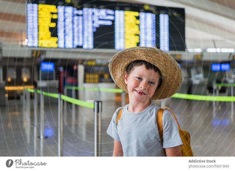 Porträt eines fröhlichen Jungen in Freizeitkleidung und Strohhut mit fehlenden Zähnen, der lächelnd in die Kamera schaut und einen Rucksack trägt, während er im Flughafen-Terminal wartet