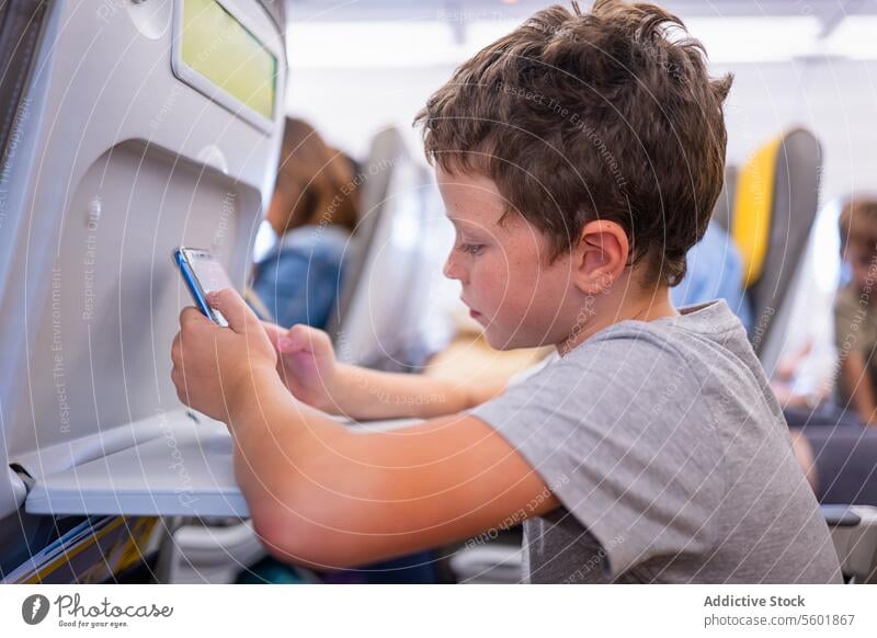 Seitenansicht eines ernsthaften Jungen in Freizeitkleidung, der ein Mobiltelefon benutzt, während er auf einem Sitz in einem modernen Verkehrsflugzeug sitzt