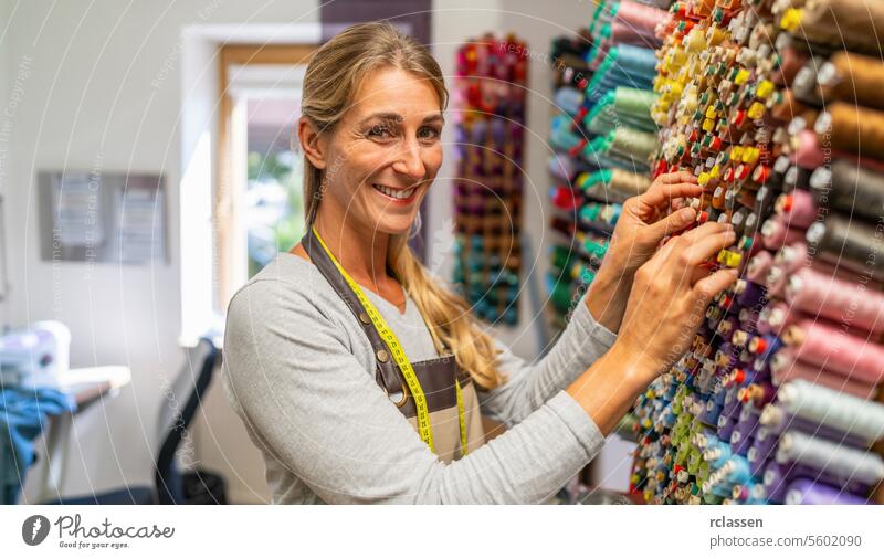 Fröhliche Näherin mit einem Maßband um den Hals wählt Fäden für eine Nähmaschine aus einem farbenfrohen, an der Wand befestigten Fadendisplay in einem Näharbeitsraum aus