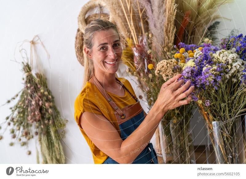 Fröhliche Floristin in senffarbenem Hemd und Jeansschürze hält einen Strauß getrockneter Blumen, umgeben von einer Reihe von Blumen vor weißem Hintergrund Kunde