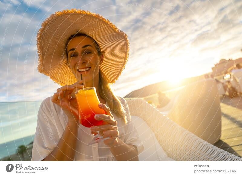 Frau mit Strohhut nippt an einem tropischen Cocktail in einem tropischen Strandhotel bei Sonnenuntergang mit unscharfem Hintergrund Paradiesstrand Playa