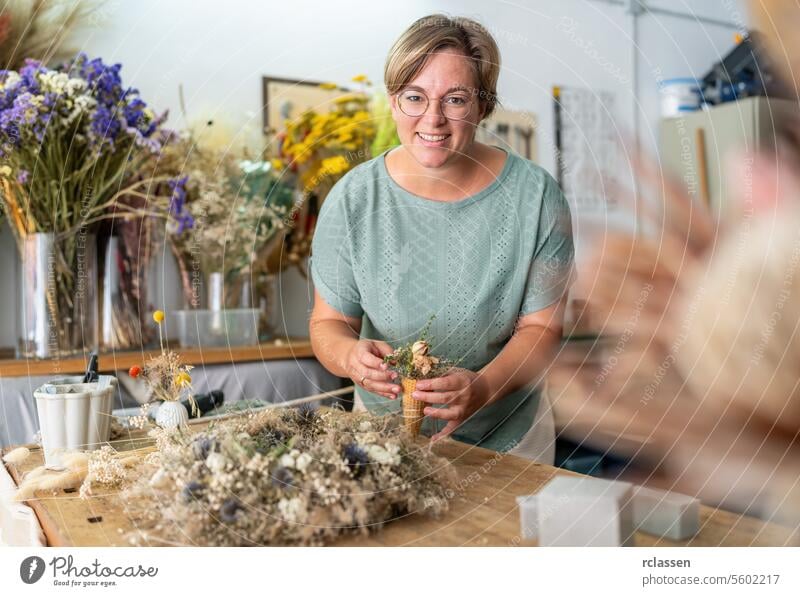 Eine fröhliche Frau in einer mintgrünen Bluse arrangiert Trockenblumen in einem Kegel, umgeben von einer lebendigen Studiokulisse mit einer vielfältigen Flora