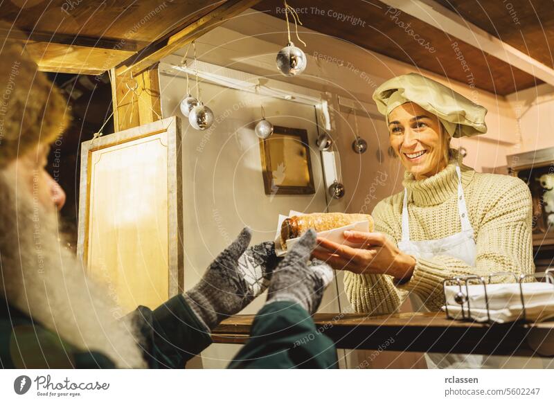 Lächelnde Verkäuferin überreicht einem alten Mann einen Baumstriezel, Trdelnik oder Kurtoscalacs an einem Stand auf dem Weihnachtsmarkt Vollbart alter Mann