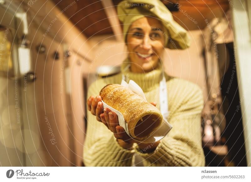 Glückliche und lächelnde Verkäuferin mit Baumstriezel, Trdelnik oder Kurtoscalacs in der Hand in einem Stand auf dem Weihnachtsmarkt Frau Lebensmittel Küche