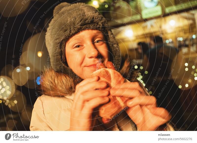 Fröhliches Kind in Winterkleidung isst eine Wurst im Brötchen auf einem Weihnachtsmarkt mit festlicher Beleuchtung freudig Frohe Weihnachten Handschuhe