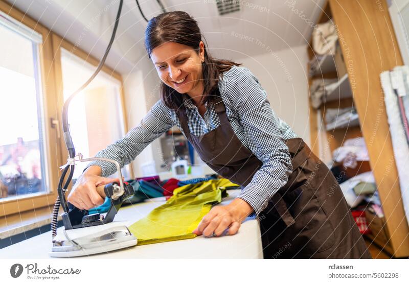 Eine lächelnde Frau bügelt ein Stück leuchtend grünen Seidenstoff in einem gut beleuchteten Nähzimmer, umgeben von Nähmaterial. Schneidern Büglerin bügelnd