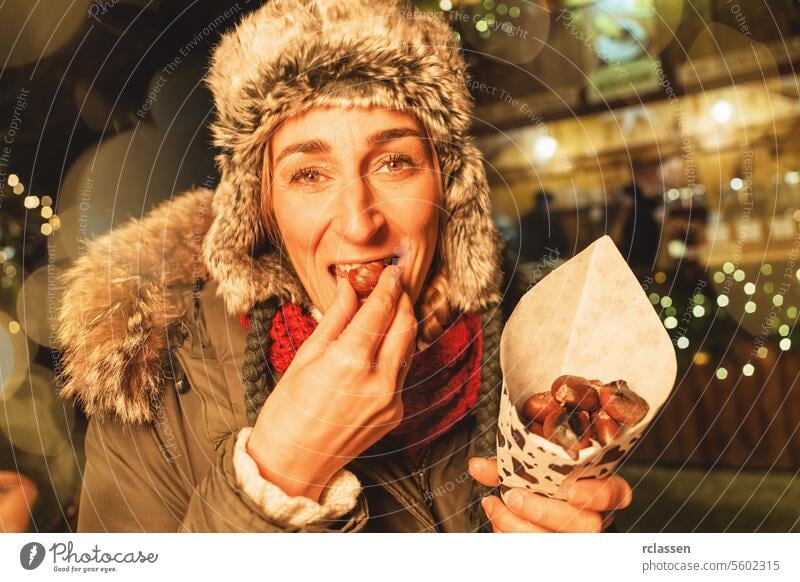 Frau isst geröstete Kastanien auf einem Weihnachtsmarkt, erfreut, mit festlichen Lichtern rundherum Frohe Weihnachten Handschuhe traditionell Glühwein Deutsch