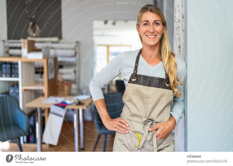 Eine fröhliche blonde Frau in einem grauen Oberteil und einer beigen Schürze steht an einem Schneiderarbeitsplatz. Sie hat ein Maßband und eine Schere in ihrer Schürzentasche und ist von organisierten Bastelmaterialien umgeben