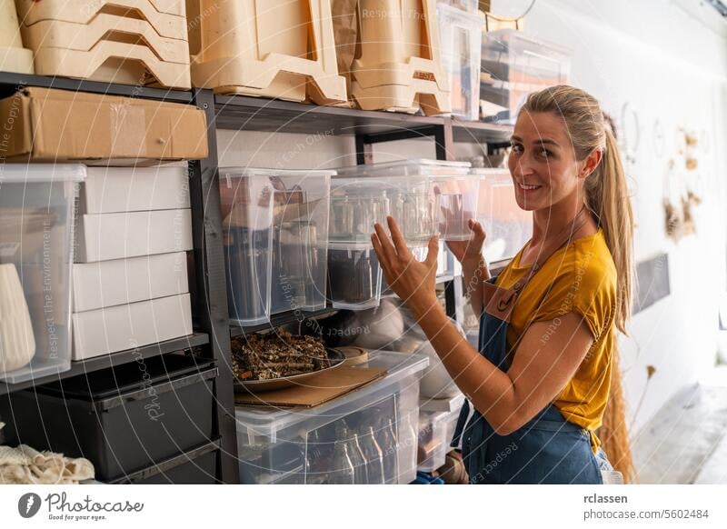 Eine Frau in senffarbenem Hemd und Jeansschürze ordnet in einer Trockenblumenwerkstatt durchsichtige Vorratsbehälter auf Regalen an und schaut lächelnd in die Kamera.