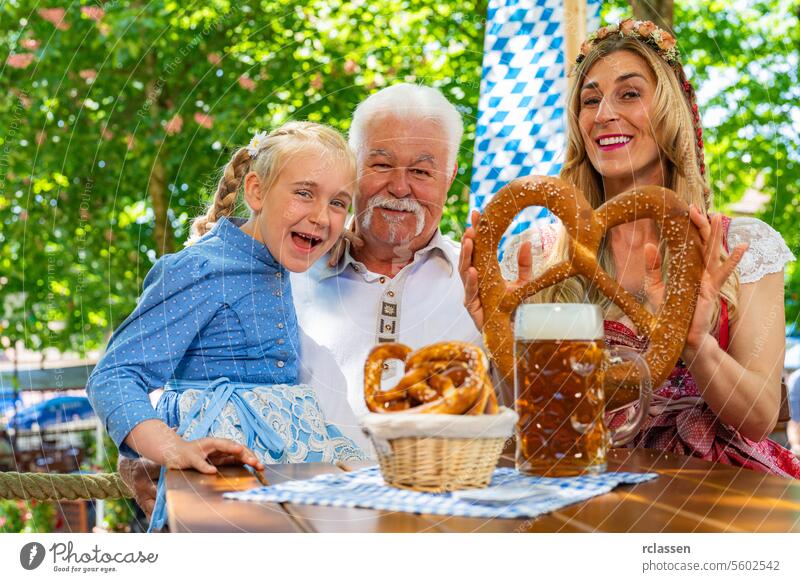 Glückliche Familie in zünftiger Tracht, die eine große Brezel hält und im bayerischen Biergarten auf dem Oktoberfest, Volks- oder Bierfest in Deutschland trinkt