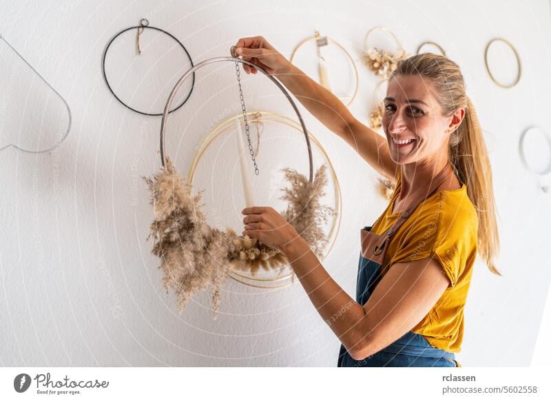 Frau lächelt, während sie eine Trockenblumen-Dekoration an die Wand hängt Lächeln Wandbehang kreatives Hobby Blumenkunst Innenarchitektur Wohnkultur Handwerk