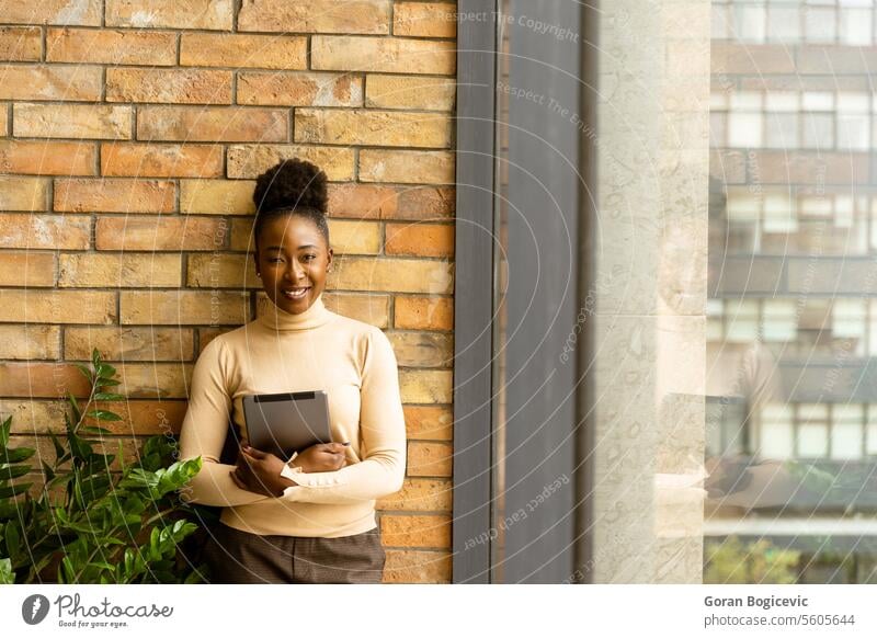 Eine junge afroamerikanische Geschäftsfrau mit digitalem Tablet steht an der Backsteinmauer in einem Büro im Industriestil. Erwachsener Afrikanisch Amerikaner