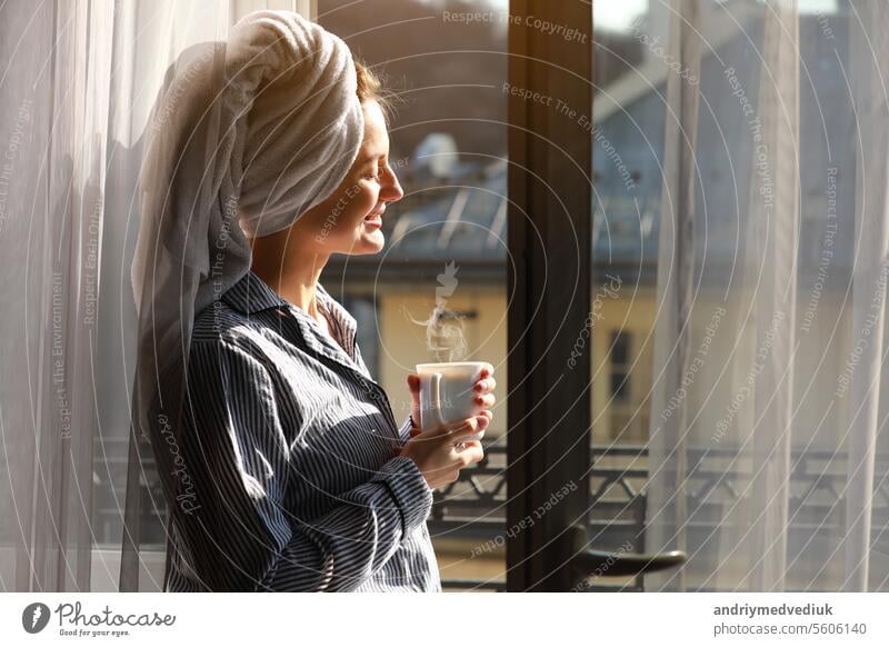 Schöne junge Frau mit Badetuch auf dem Kopf hält eine weiße Tasse mit heißem Kaffee oder Tee zu Hause am Fenster. Lächelndes Mädchen nach der Dusche genießt Aroma Getränke und haben Frühstück am Morgen