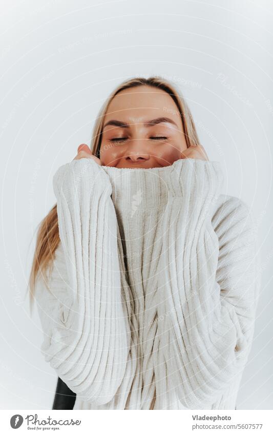 Junge blonde Frau, bekleidet mit weißem Pullover, in einen Kragen gewickelt, schaut in die Kamera, weißer Hintergrund jung Atelier Blick lieblich posierend