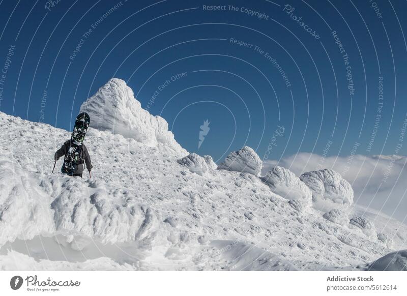 Rückenansicht eines nicht erkennbaren Snowboarders, der seine Ausrüstung auf einen schneebedeckten Berg mit einzigartigen Schneeformationen unter einem klaren blauen Himmel in Japan trägt