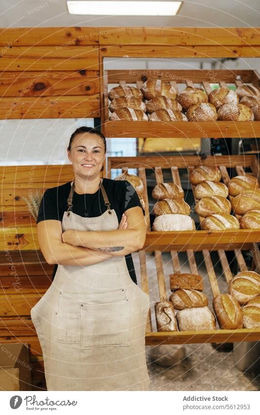 Glückliche Verkäuferin neben Brot in einer Bäckerei Regal Brotlaib selbstbewusst Lächeln die Arme verschränkt Schürze frisch gebacken Lebensmittel hölzern