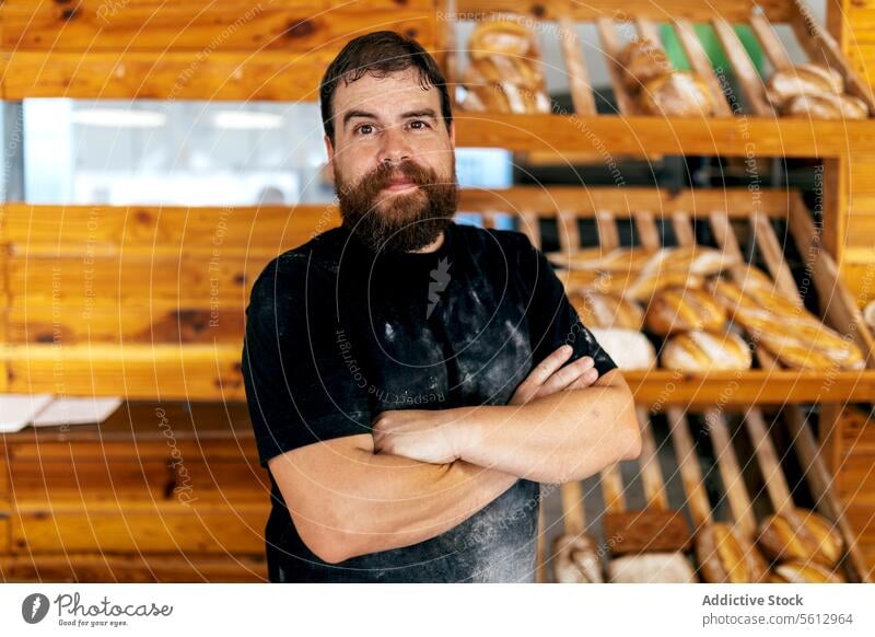 Ruhiger Verkäufer neben Brot in einer Bäckerei Mann selbstbewusst Regal die Arme verschränkt Laden Werkstatt Unschärfe Brotlaib Vollbart Freizeitkleidung frisch