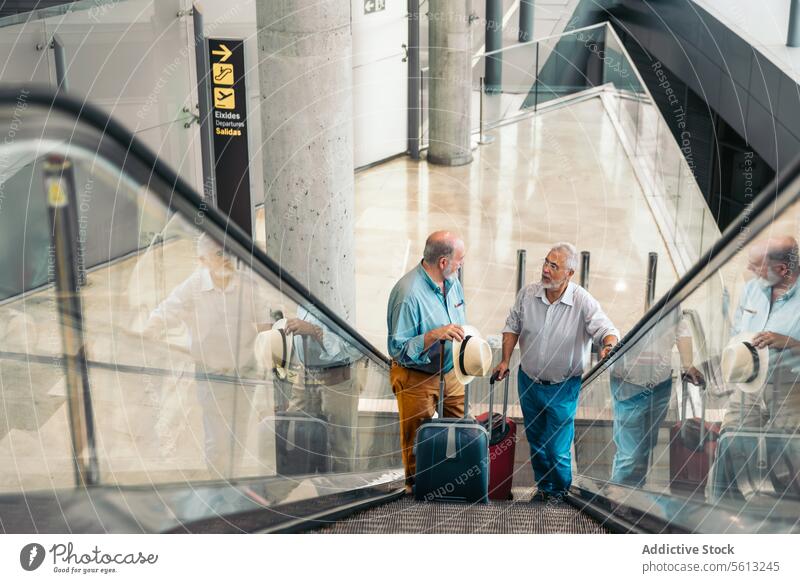 Freunde mit Gepäck auf der Rolltreppe im Flughafen reden Reisender Männer Terminal hoher Winkel stehen bewegend Treppe Koffer Hut Transit Senior Urlaub