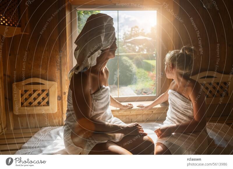 Rückenansicht einer Mutter und ihrer Tochter in einer finnischen Sauna, mit Blick aus dem Fenster, eingewickelt in Handtücher in einem Wellness-Hotel Bademantel