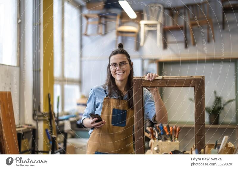 Porträt einer jungen Schreinerin, die in ihrer Werkstatt ein Mobiltelefon benutzt Möbel Zimmerer wiederherstellend Schreinerei Holz Stuhl Gebäude Herstellung