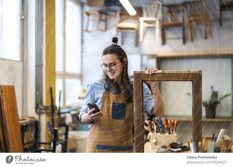 Porträt einer jungen Schreinerin, die in ihrer Werkstatt ein Mobiltelefon benutzt Möbel Zimmerer wiederherstellend Schreinerei Holz Stuhl Gebäude Herstellung