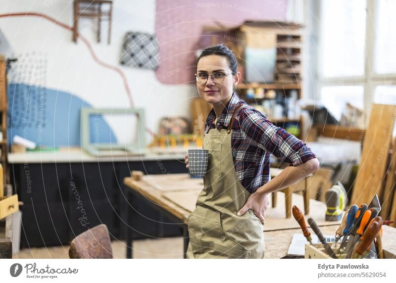 Porträt einer jungen Schreinerin mit einer Tasse Kaffee in der Hand in ihrer Werkstatt Möbel Zimmerer wiederherstellend Schreinerei Holz Stuhl Gebäude