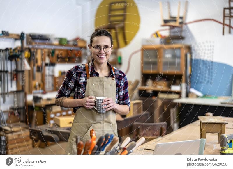 Porträt einer jungen Schreinerin mit einer Tasse Kaffee in der Hand in ihrer Werkstatt Möbel Zimmerer wiederherstellend Schreinerei Holz Stuhl Gebäude