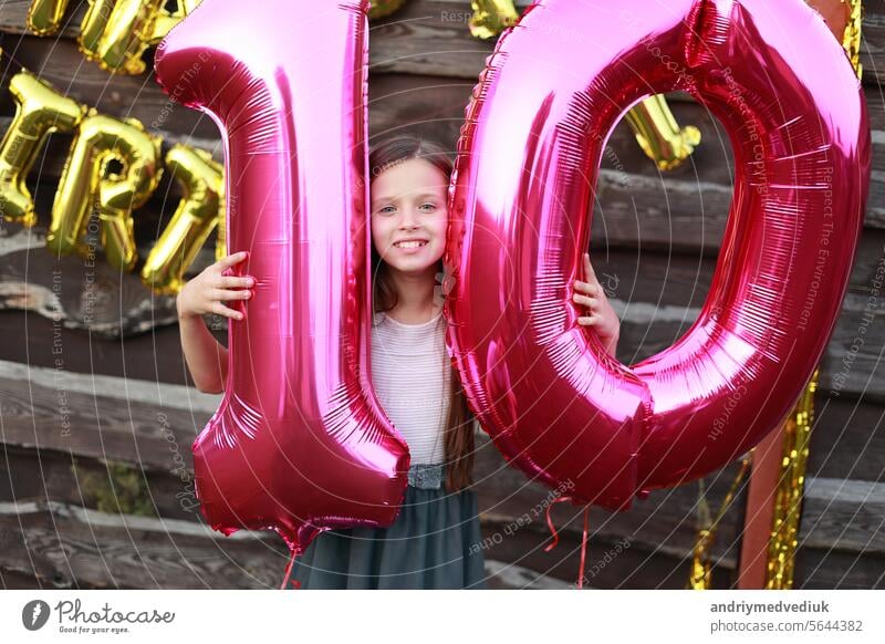 Fröhlich kleines Mädchen hält zwei große rosa Folienballons, Nummer 10 im Freien auf hölzernen Wand Hintergrund mit goldenen Ballons Aufschrift alles Gute zum Geburtstag. Zehn Jahre Jahrestag Feier Partei zu Hause